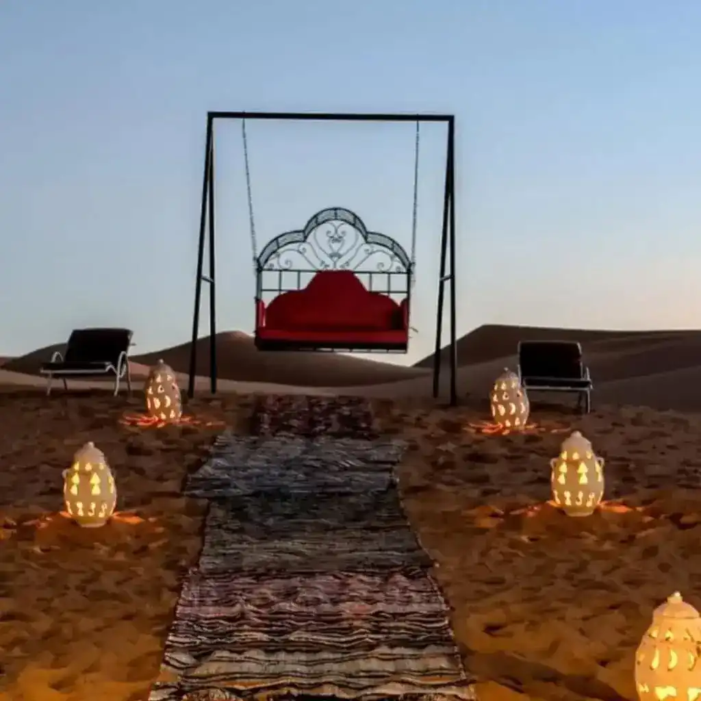 A cushioned swing and glowing lanterns set on the sand at a desert camp for a private dinner